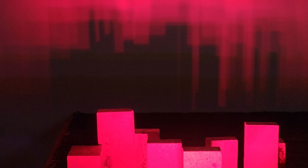 Picture of sculpture showing blocks illuminated with red light, casting a skyline shadow on a wall