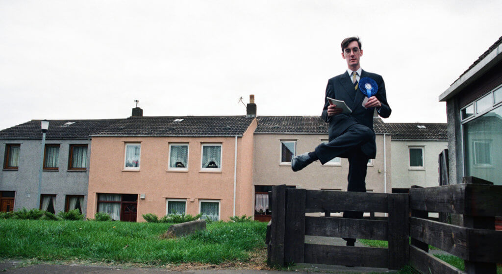 Jacob Rees Mogg stepping over a garden fence on a housing estate whilst campaigning as Tory candidate in Central Fife, 1997. Painted terraced housing in background. Photo by Colin McPherson.