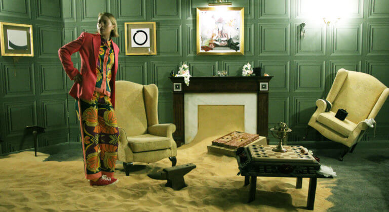 A woman stands in a green-panelled drawing room. She is wearing a brightly coloured pyjama-suit, and has her hand on her hip. There are drifts of sand on the floor, and furniture and other objects at strange angles.