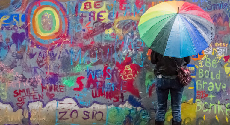 A woman with her back to camera is holding a rainbow umbrella and facing a wall covered in brightly-coloured graffiti.