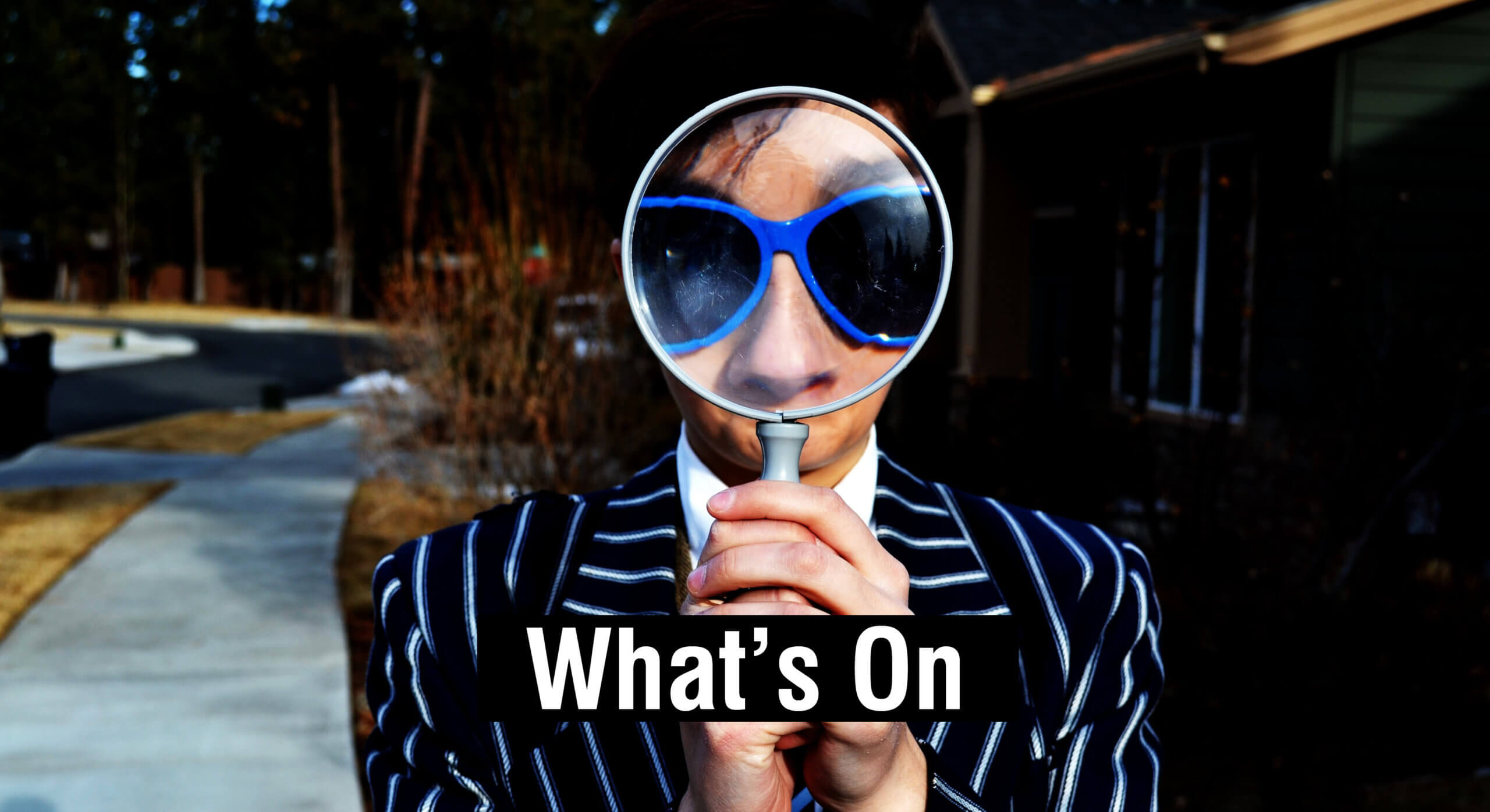 Figure in pinstripe suit wearing blue sunglasses. Figure is hold a magnifying glass over their face filling the glass with just blue sunglasses.