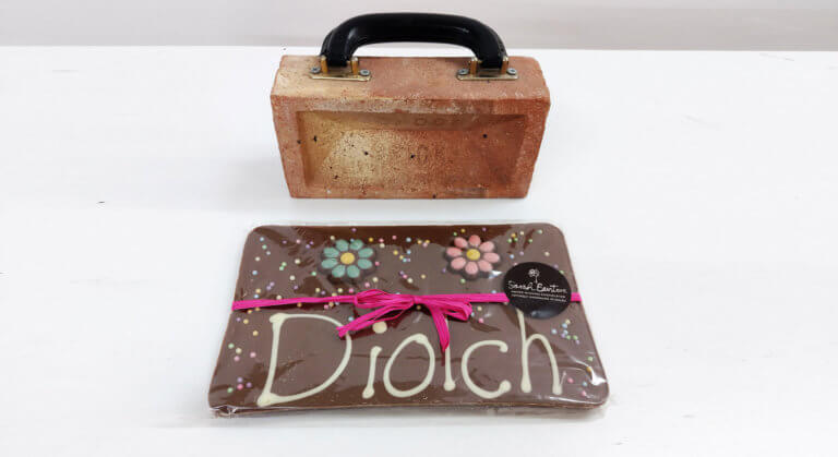 A large slab of chocolate with the word 'Diolch' on it is sitting on a table in front of a brick.