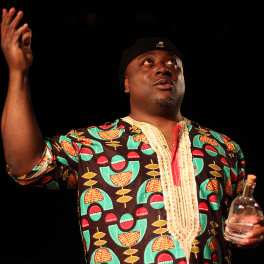 A picture of the poet Eric Ngalle Charles. He is wearing a traditional colourful Cameroonian shirt and a black Kangol cap. He is looking upwards, his right hand raised a a glass bottle of water in his left.