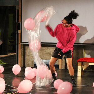A young woman wearing a pink hoodie and pink sunglasses is shaking a bag full of pink balloons onto a a stage.