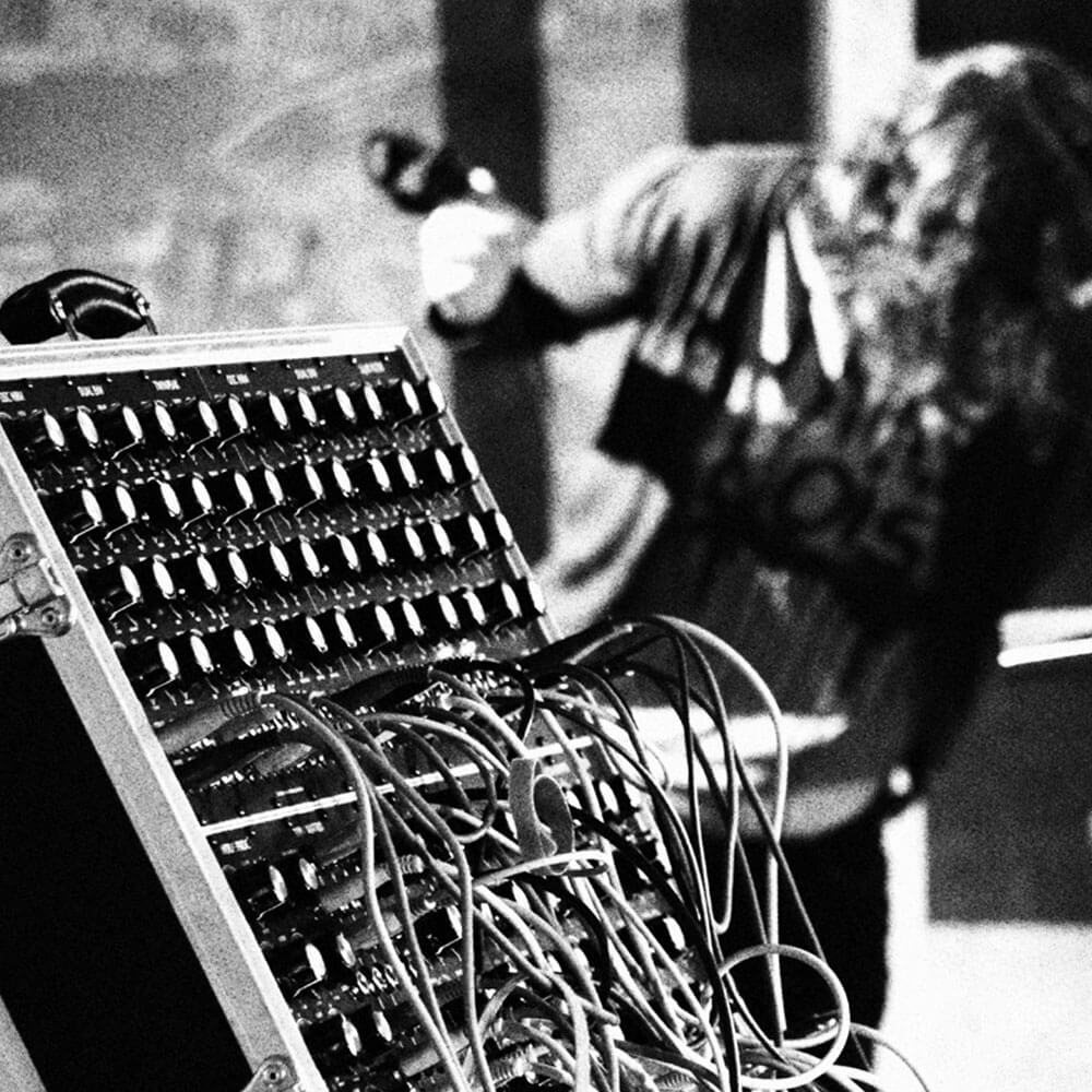 Black and white image of a sound mixer in foreground. A musician is playing an electronic instrument in the background.