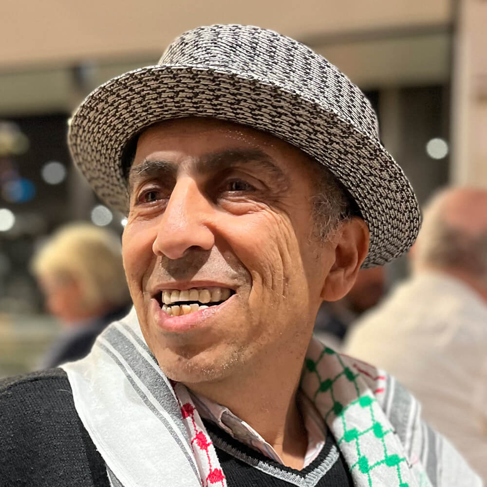 Prof Mazin Qumsiyeh. He wears a boater hat and a scarf or keffiyeh with a Palestinian design.