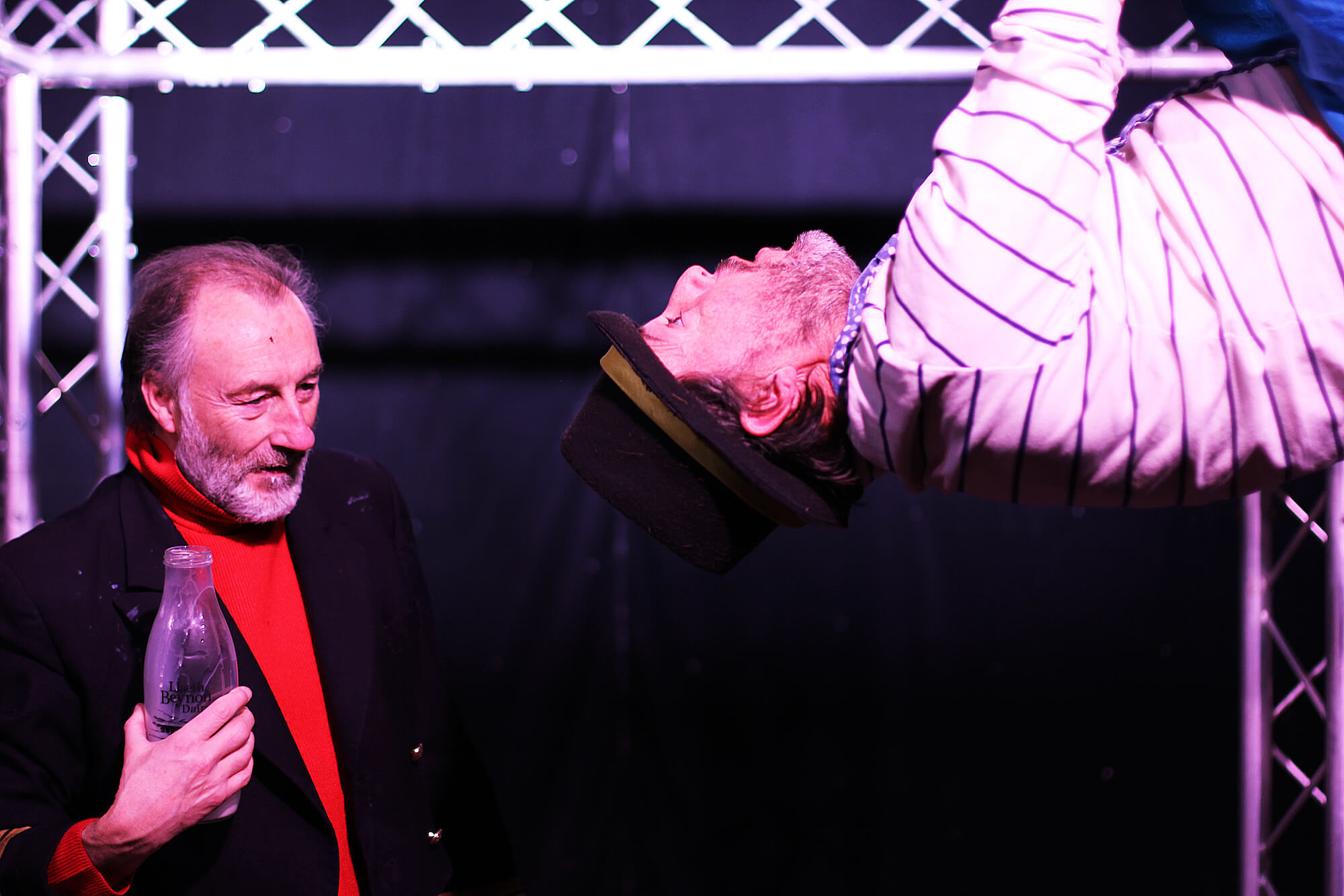 Two men onstage talking. One is holding a milk bottle. The other is hanging upside down.