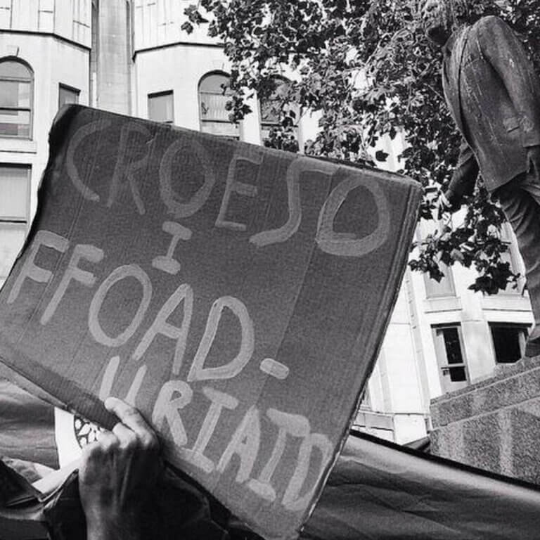 A black and white photo of a protestor holding a sign reading "refugees welcome" in Welsh, beneath the statue of Aneurin Bevan in Cardiff.