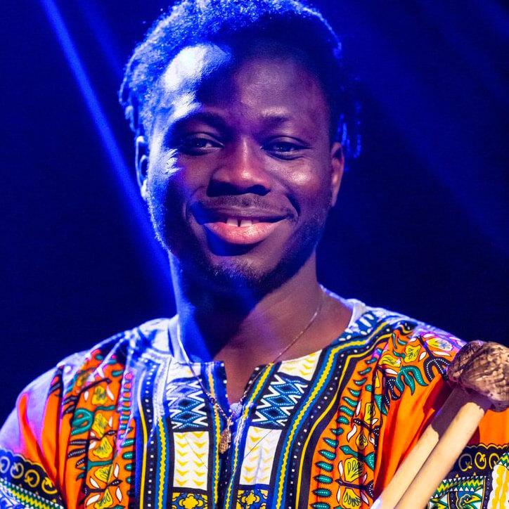 Portrait of N'famady Kouyate. He is wearing a brightly coloured dashiki and holding balafon mallets.