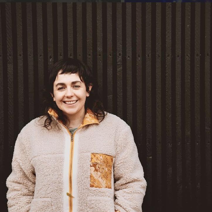 Portrait of Rhiannon Mair. She is wearing a fleece jacket and standing in front of a corrugated grey wall.