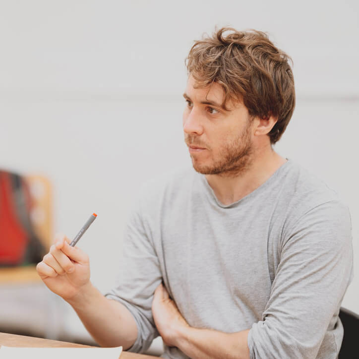 Image of Steffan Phillips. He is sitting at a table wearing a grey sweater and holding a pen.
