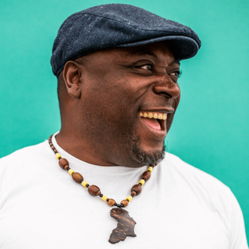 A portrait of Eric Ngalle Charles. He is wearing a white t shirt and blue cap, and a necklace with a map of Africa.